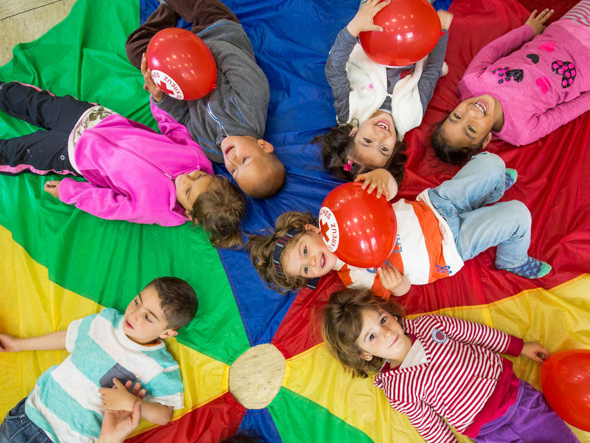 viele Kinder liegen im Kreis auf einem bunten Schwungtuch und lachen in die Kamera über Ihnen